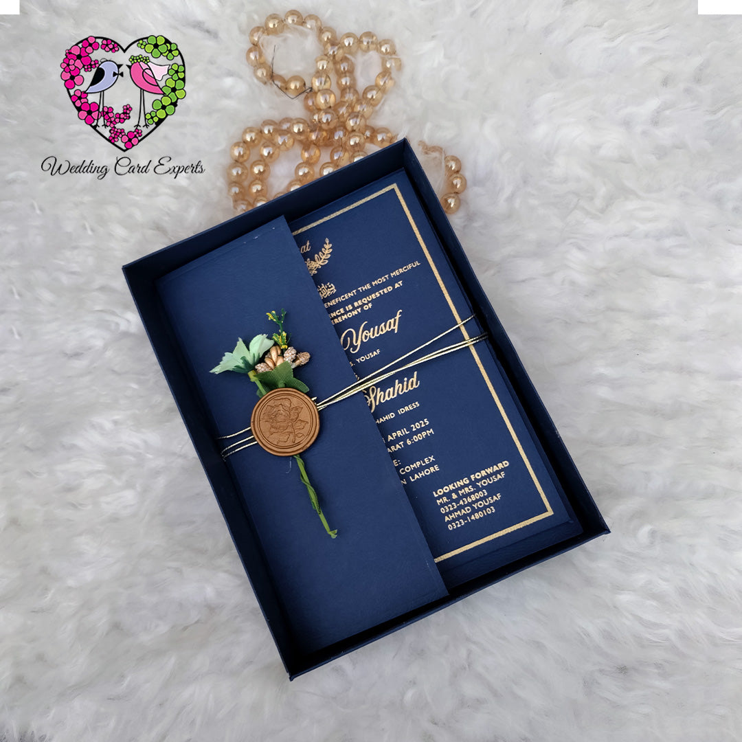 Navy blue wedding invitation with gold text and a decorative brooch on a textured white surface.