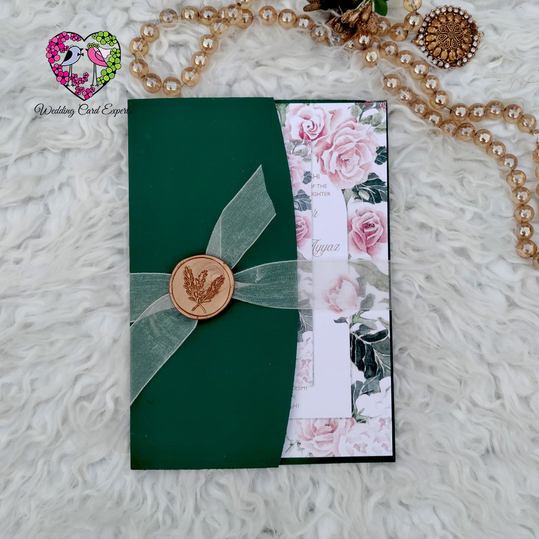 Pakistani wedding invitation card — hunter green pocket folder tied with sheer sage organza ribbon and copper botanical wax seal, blush floral card peeking out, wedding cards from Pakistan