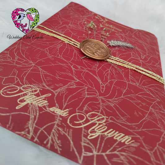 Pakistani wedding card close-up — gold foil botanical print crimson red envelope with copper wax seal and double gold twine wrap, wedding card Pakistan luxury