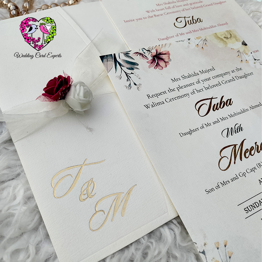 Pakistani Wedding invitation card with floral design and foil-printed monogram on a textured surface.